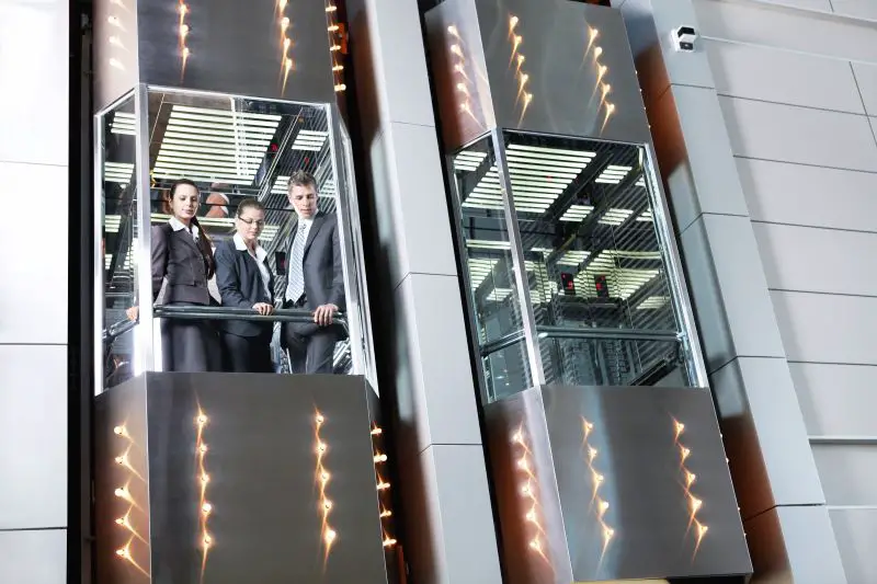 Three business professionals standing inside a glass elevator in a modern office building.