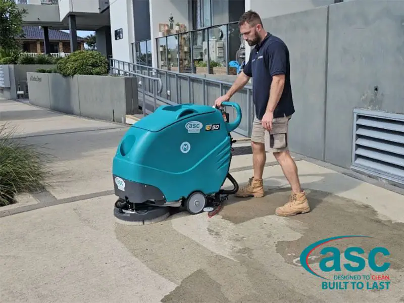 Man using a commercial floor scrubber machine to clean outdoor concrete surface at a commercial building entrance.