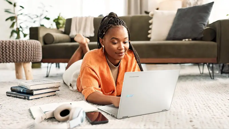 Woman using laptop while lying on the floor at home, with books, headphones, and smartphone nearby