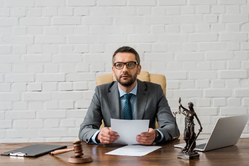 Serious bearded lawyer with eyeglasses in a suit sitting at the workplace holding documents and looking at the camera.