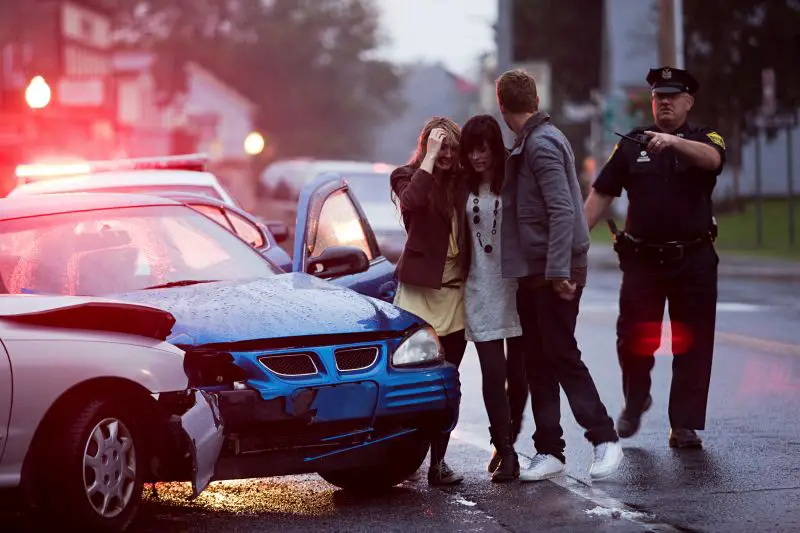 Woman comforted by man at the scene of a car accident as a police officer directs traffic.