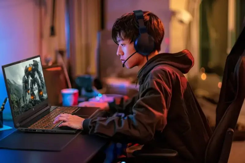 An Asian boy wearing a headset is playing games on a laptop.