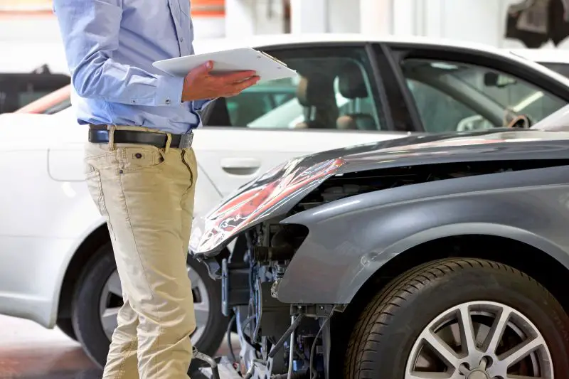 Insurance Assessor With A Clipboard Inspecting A Damaged Vehicle At The Garage.