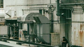 Industrial building exterior with large ventilation ducts and a person sitting on a ledge taking a break