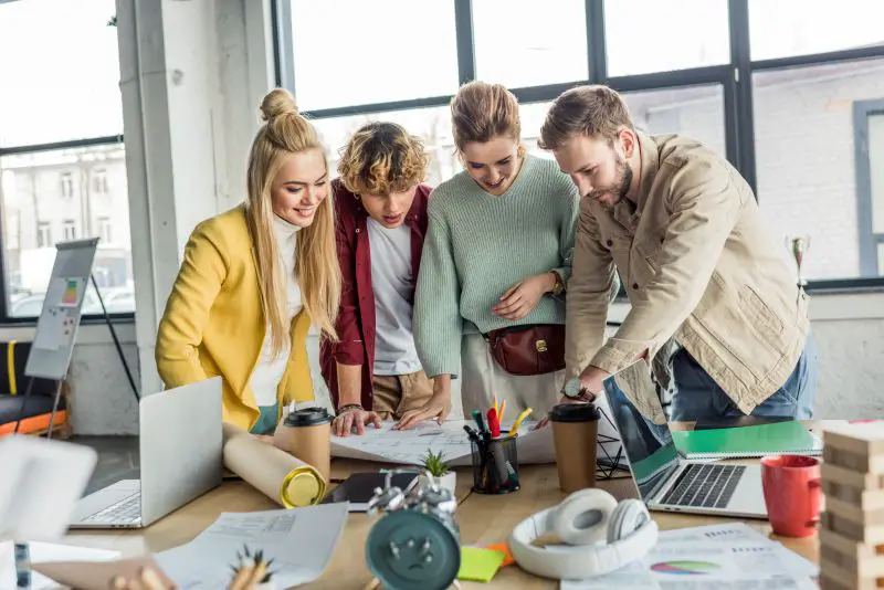 group of female and male architects working on blueprint at desk with laptops in loft office