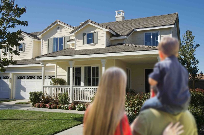 Family viewed from behind standing on a lawn and looking toward a large suburban house, symbolizing home ownership, stability, and protecting family assets.