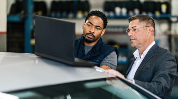 Male customer and African American car mechanic using Garage Management Systems software on laptop in auto repair shop.