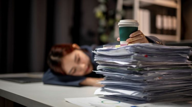 A woman is sleeping on a desk with a pile of papers and a cup of coffee. The scene suggests that she is working late and is exhausted