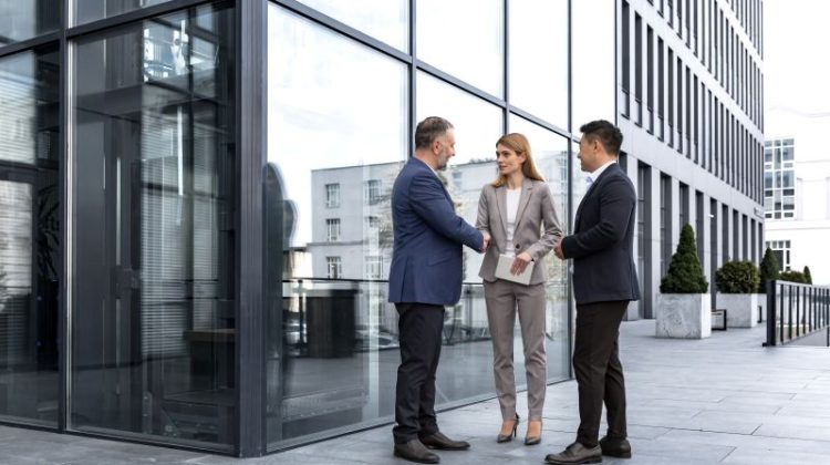 Three business professionals in modern business attire are gathered for a handshake outdoors in front of a modern office building.
