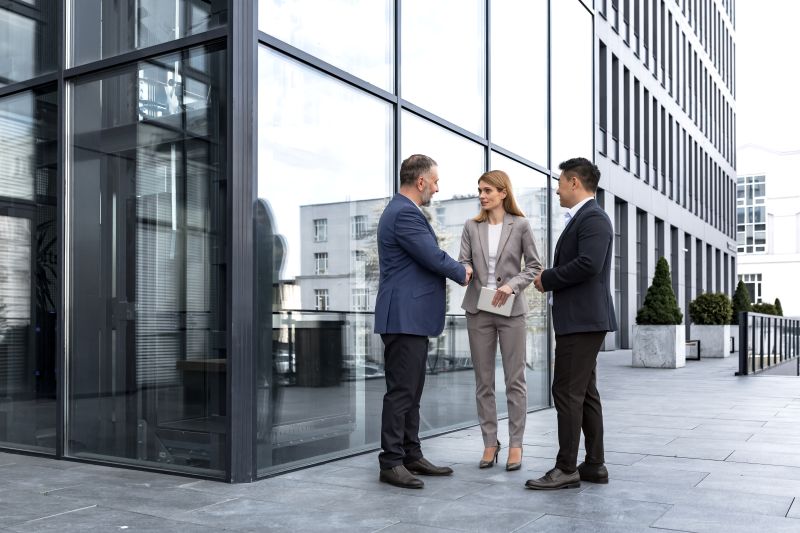 Three business professionals in modern business attire are gathered for a handshake outdoors in front of a modern office building.