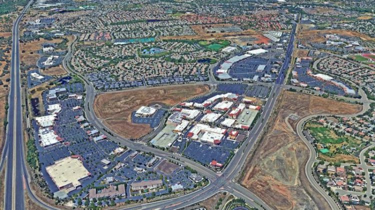 Close-up of shallow, muddy water with green plant stems emerging from the surface, possibly in a wetland or flooded area, partially obscured by a blurred foreground.