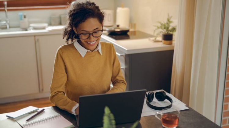 A smiling woman sits at a dark table in a sunny kitchen and looks down at the laptop in front of her.