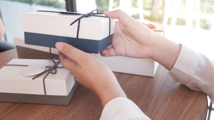 Two hands holding a white and blue gift box wrapped with a thin cord. The box is being held over a light brown, circular wooden table.