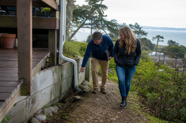 Two people inspect the exterior of a house while walking along a pathway beside the property.