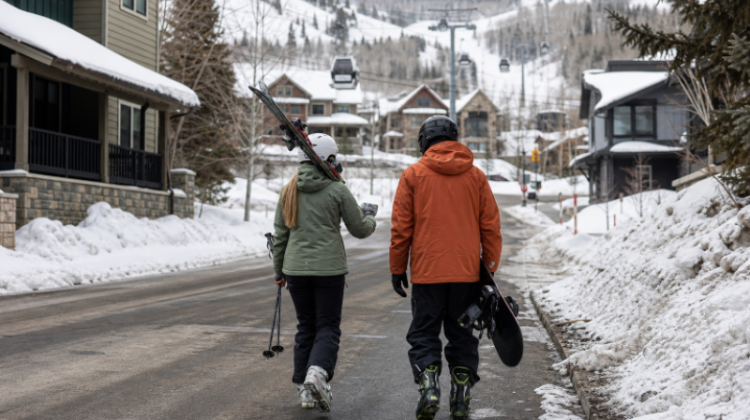 Two skiers walking through a snow-covered Park City neighbourhood toward mountain lifts and gondolas.