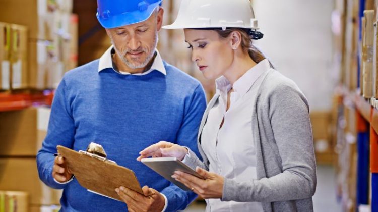 Two warehouse professionals comparing product information on a clipboard and tablet while researching customer terminology and B2B markets.