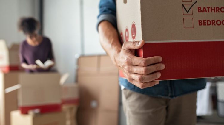 Person holding a cardboard moving box while another person packs boxes in the background.