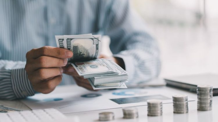 Person counting US dollar banknotes beside financial documents and ascending stacks of coins.